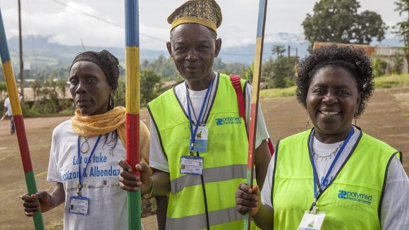 Two women and a man are carrying measuring sticks to calculate the dosage of Mectizan. Two of them are wearing high-vis tops.