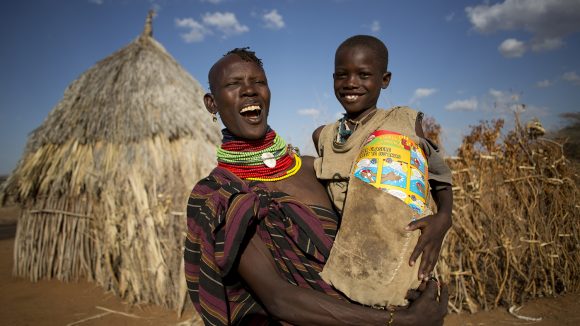 A woman holding a child, smiling and standing outside a thatched dwelling.
