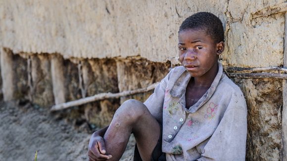 Nananga sits on the floor holding one knee and looking at the camera.