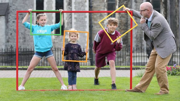 Three children hold picture frames up to their faces and a judge of the Junior Painter Awards is looking at them with his hand on his chin.