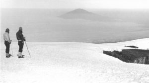 An old black and white photo shows two people standing on a vast mountain landscape covered in snow.