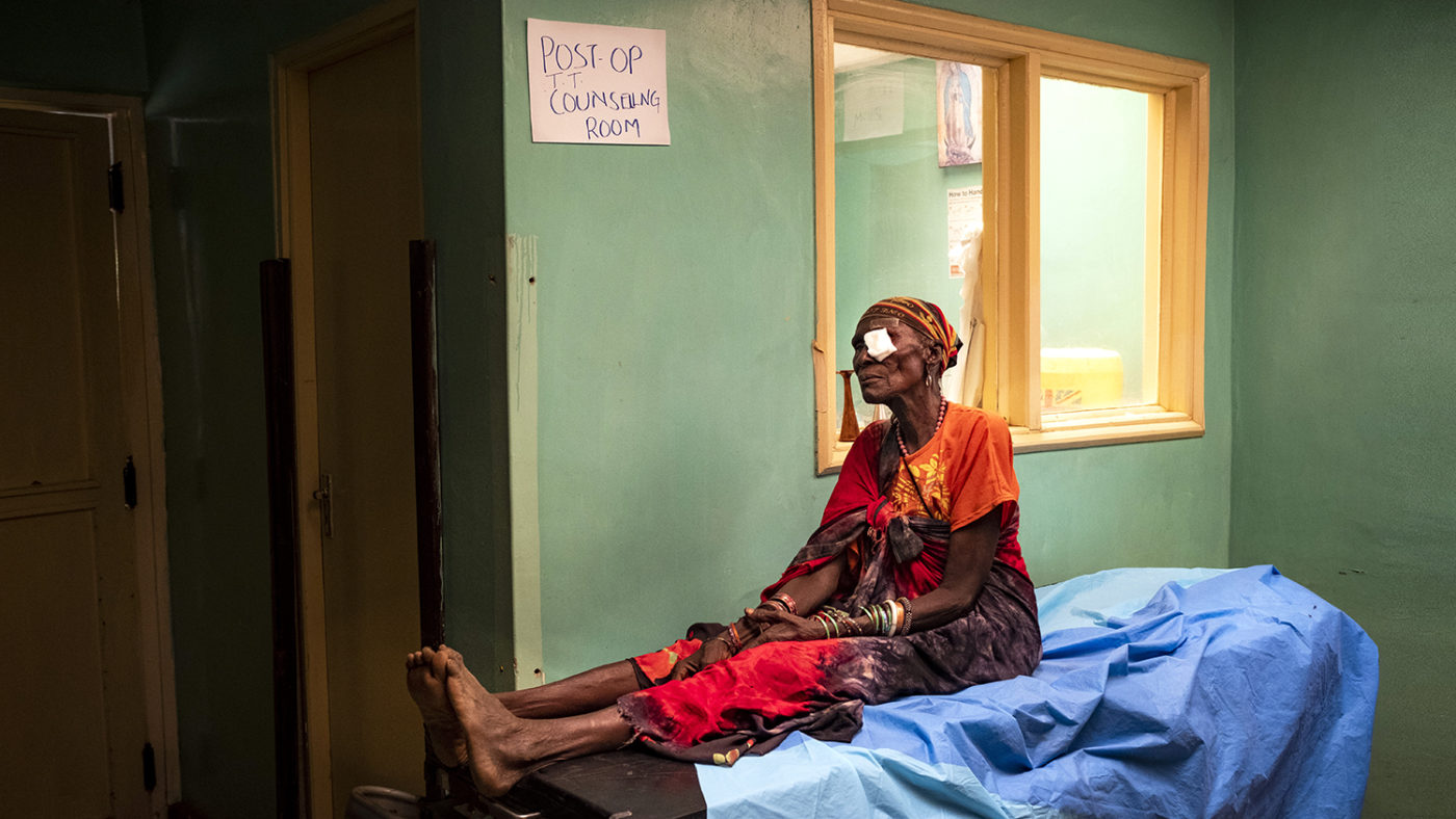 A lady sits on a bed with a eye patch over one eye.