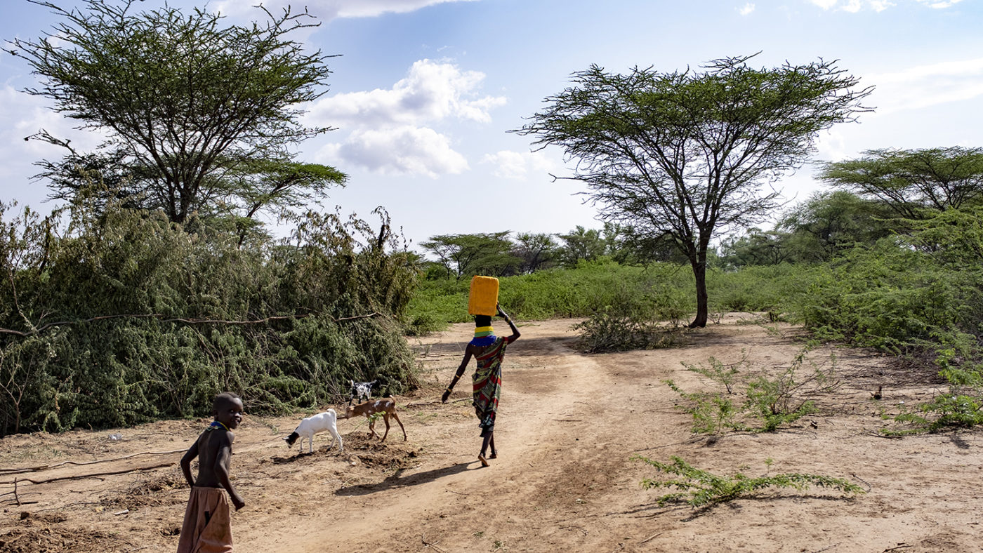 A women walks with a plastic container on her head, a child follows her.
