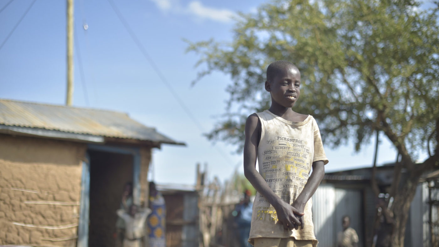 A young boy stands outside in the sun wearing light coloured clothes. There is a small house to the left and a tree to the right.