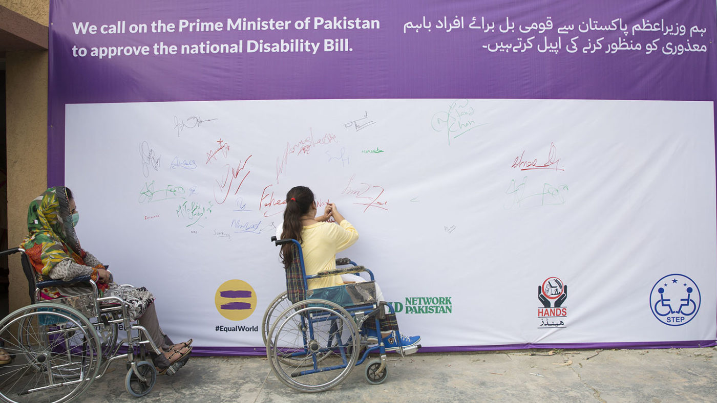 Two women in wheelchairs sign a petition on a wall.