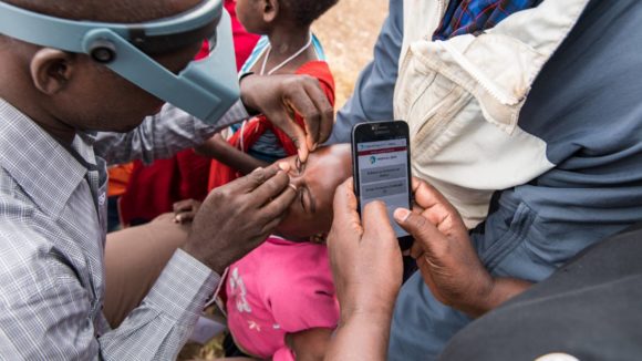 A Tropical Data trainee examines a young child for trachoma while the recorder enters data into a mobile phone.