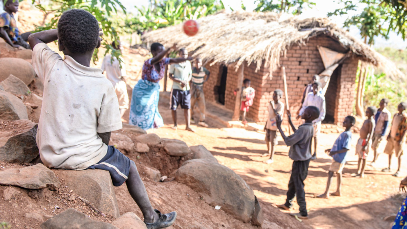 Taonga sits on the rocks shielding his eyes, a people play with a ball in the distance.