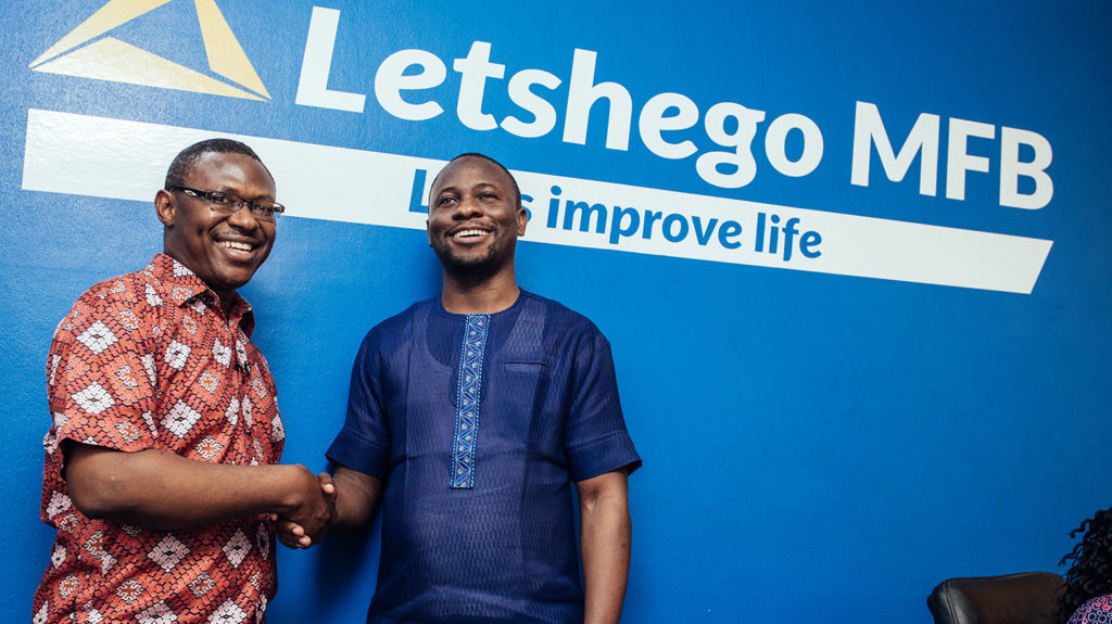 Two men shake hands in front of a sign for Letshego bank.