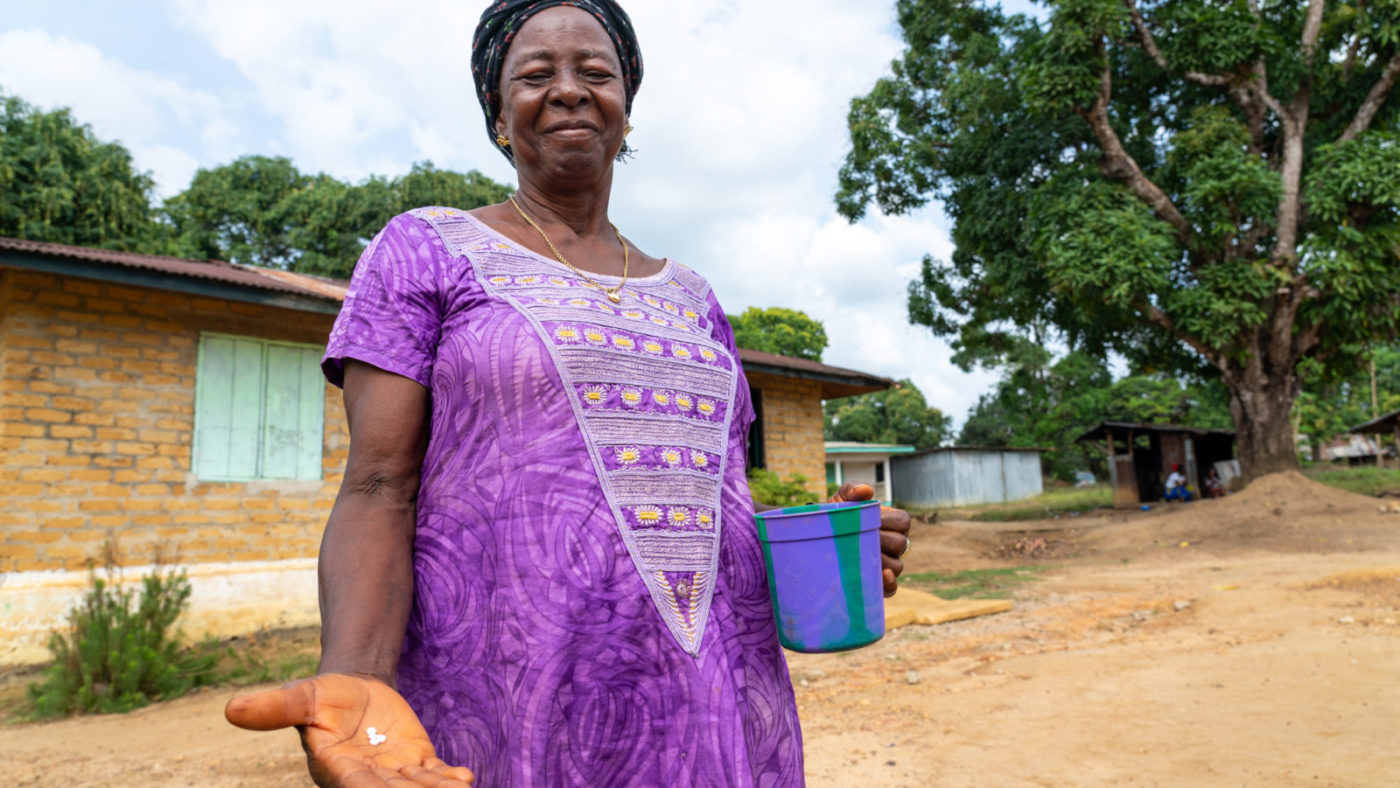 A woman stands outside, holding out her hand to show medication and holding a cup in her other hand.
