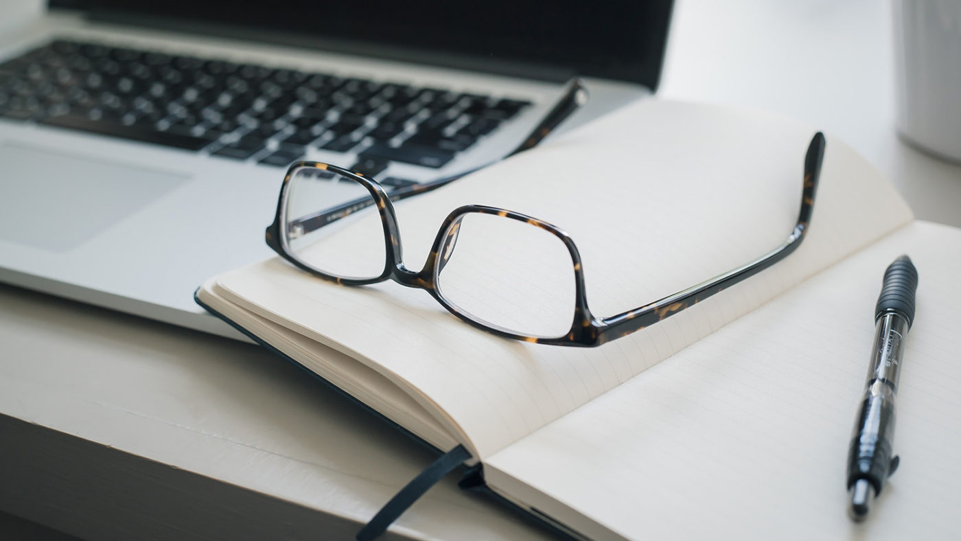A pair of glasses, a pen and a computer on a desk.
