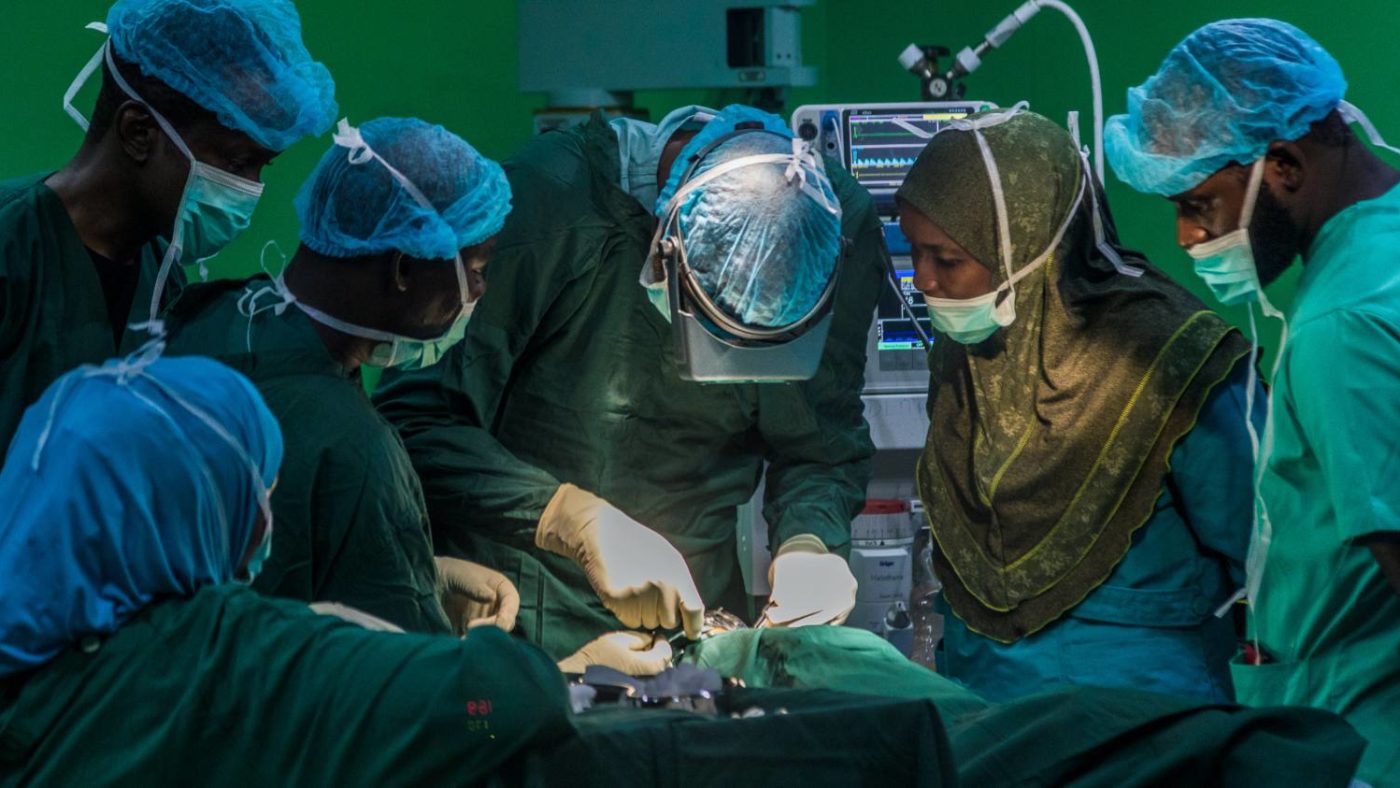 Muzi undergoing surgery in an operating theatre, surrounded by five medical professionals wearing gowns and masks.