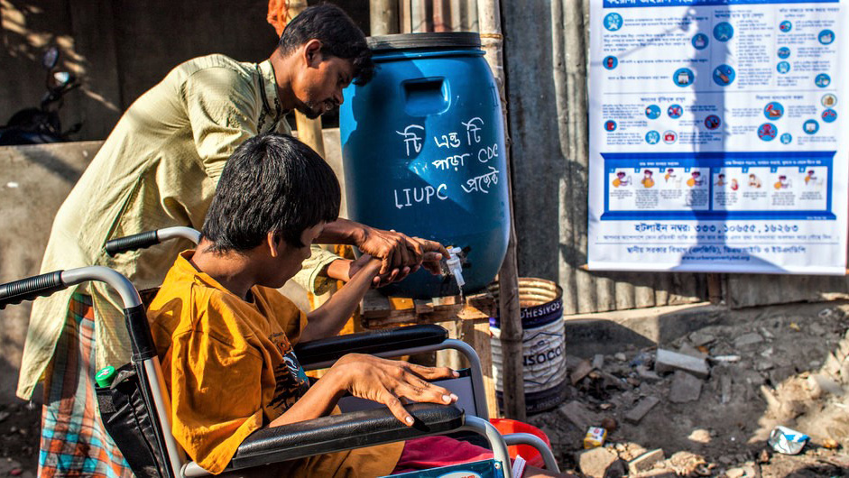 A man who uses a wheelchair is assisted by another man to use a water tap to wash his hands.