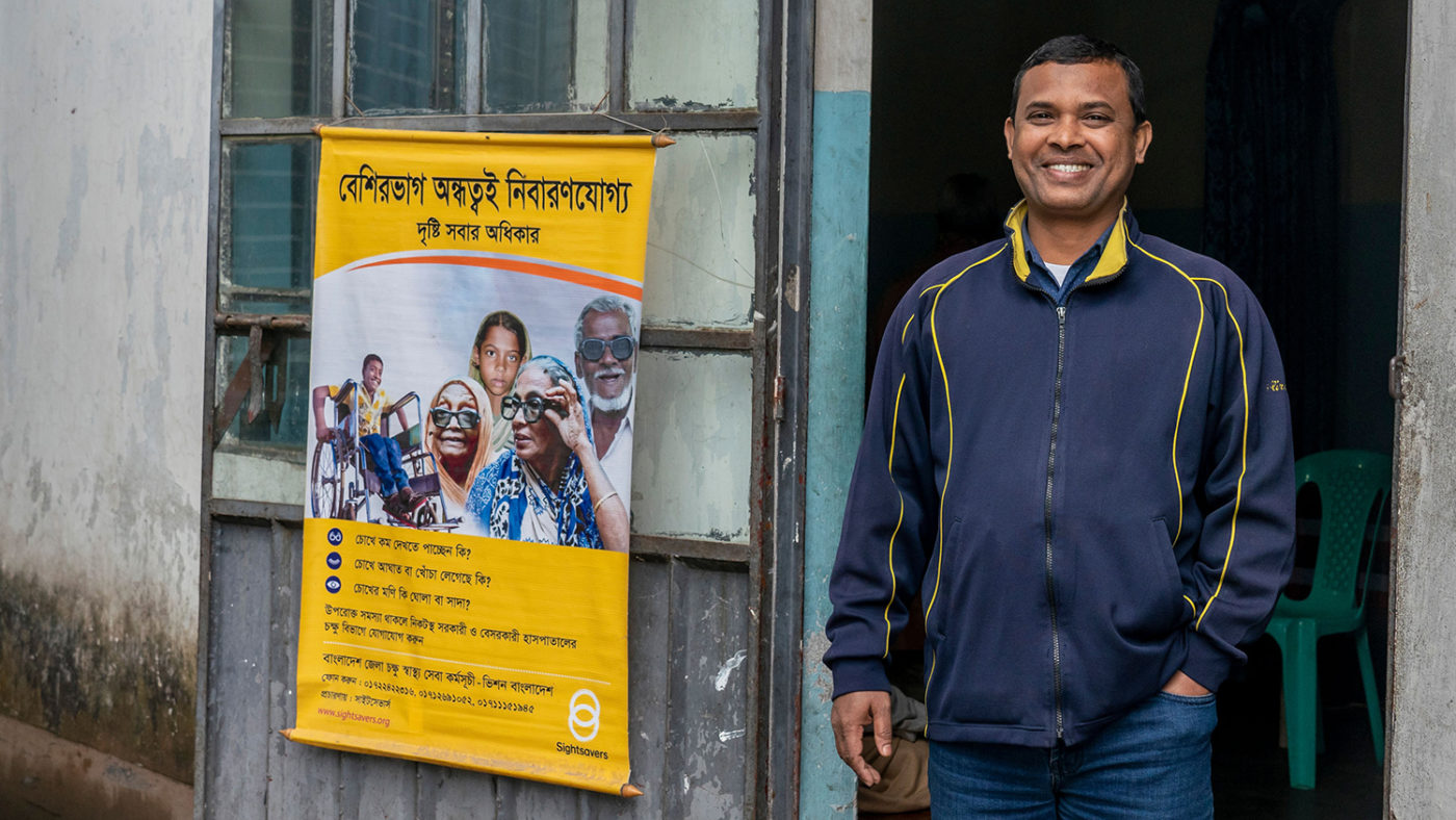 A man standing smiling, next to a Sightsavers' sign in Bangla.