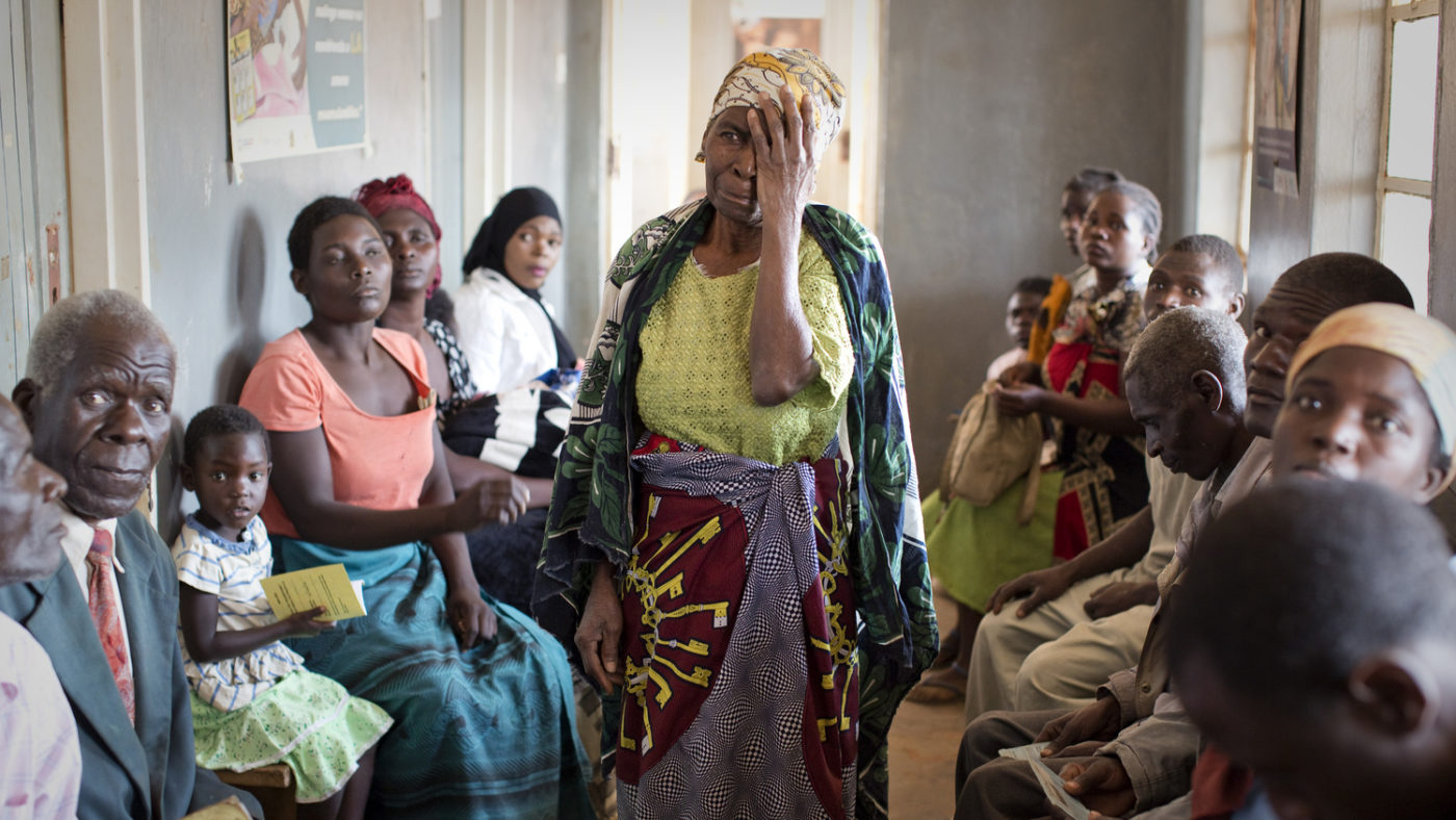 A woman standing in a roomful of other people. She is covering one eye with her hand.