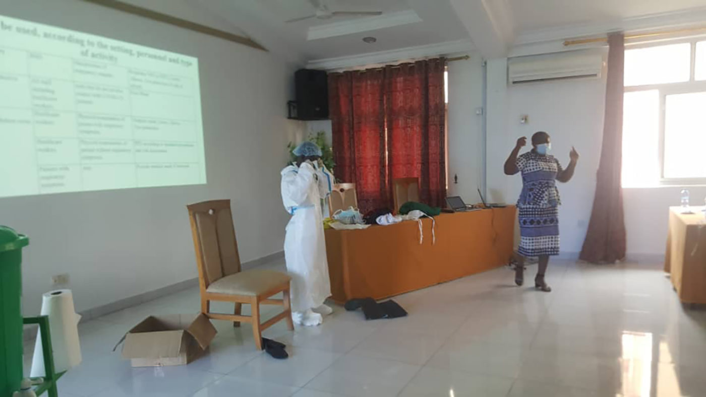 two people stand at the front of a classroom. Both are wearing masks and gowns.