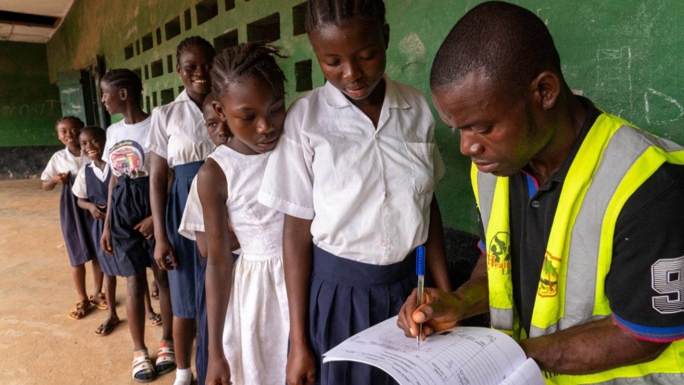 A man writes in a book while children queue up next to him.