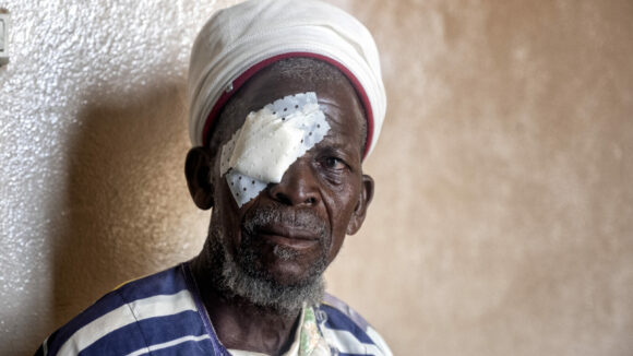 A man sits against a wall after having trachoma surgery. He has a bandage covering one eye.