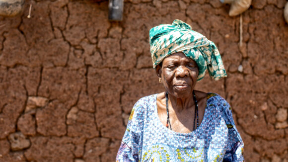 An elderly lady in colourful traditional clothing stands in front of a wall.