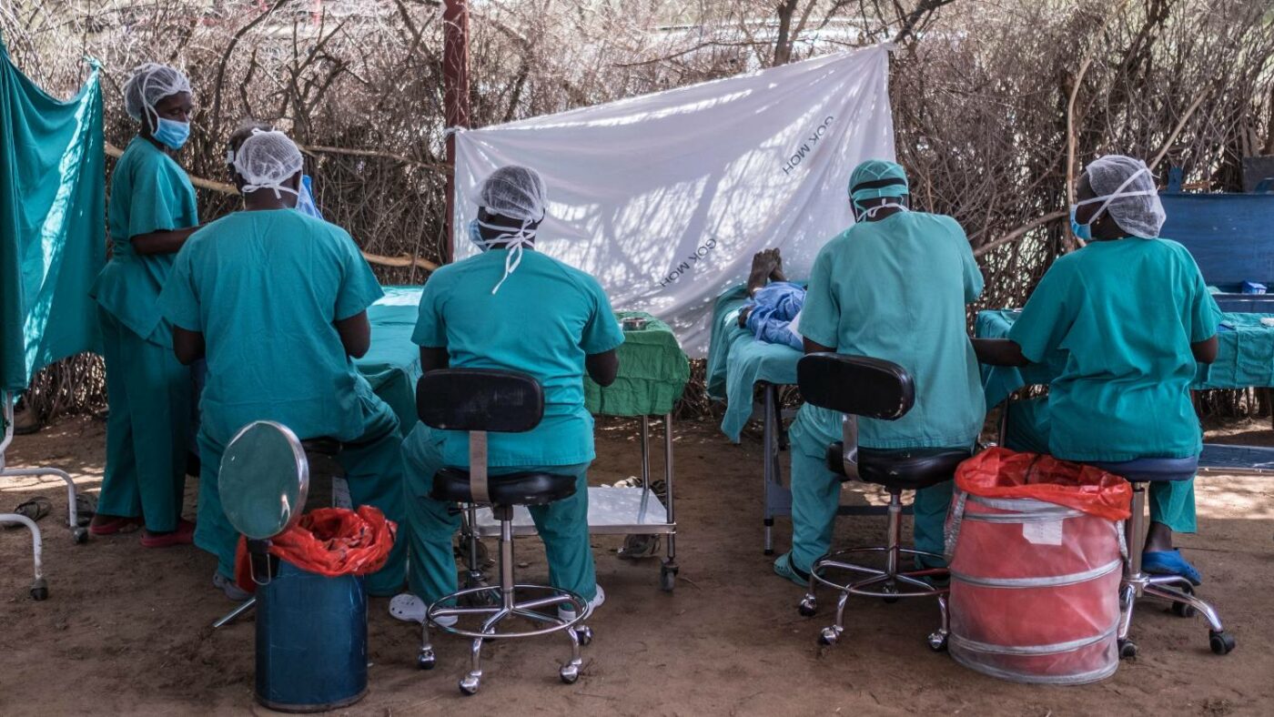 Five health workers wearing scrubs and masks attend to two trachoma patients. Sticks are used to demarcate the surgery.