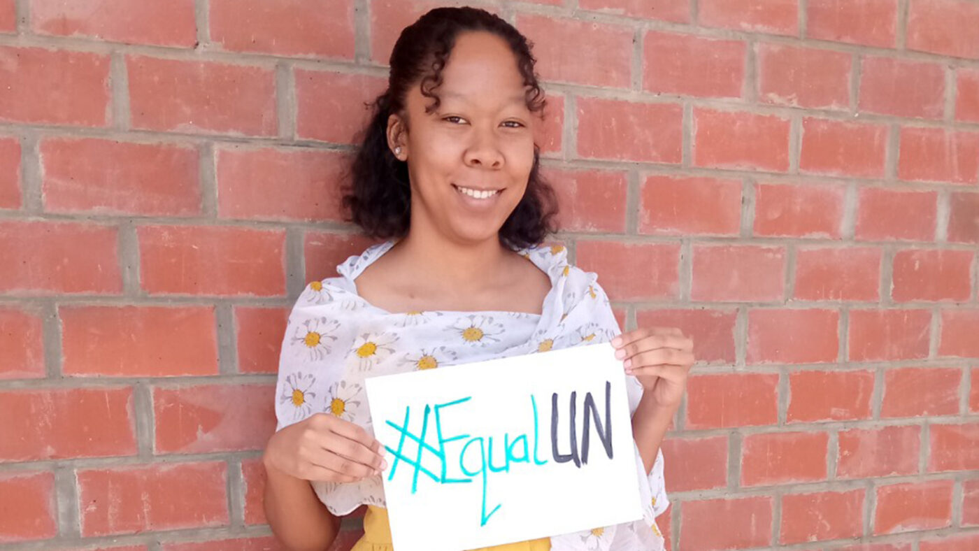 A woman standing by a brick wall holds a sign reading #EqualUN.