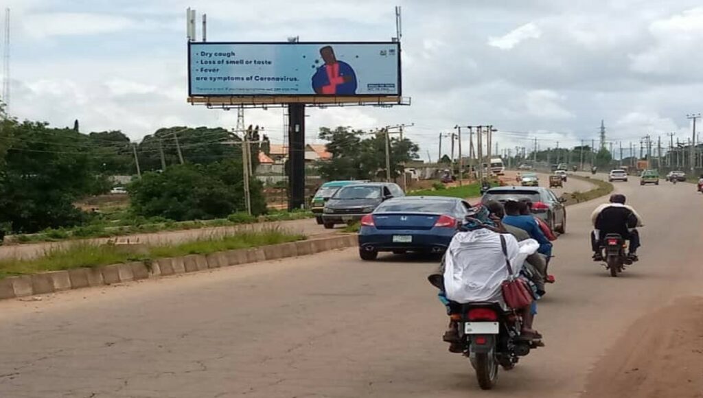 A billboard with COVID-19 messaging on it can be seen at the side of a road with cars a bikes going along it.