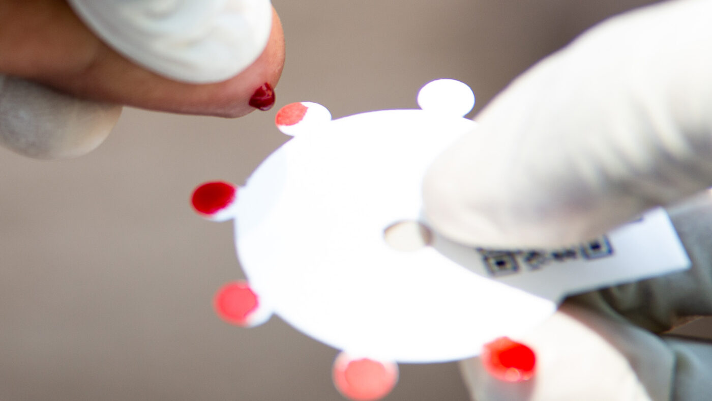 A drop of blood falls from someone's finger onto filter paper.