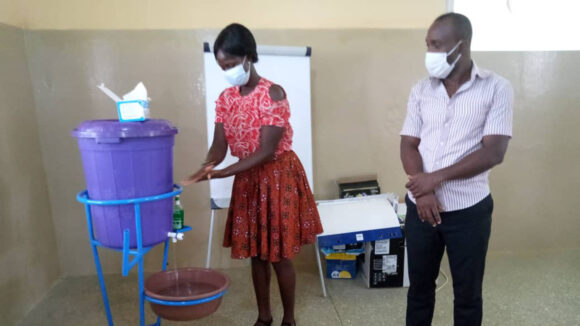 A woman washes her hands during COVID-19 response training while a man observes.
