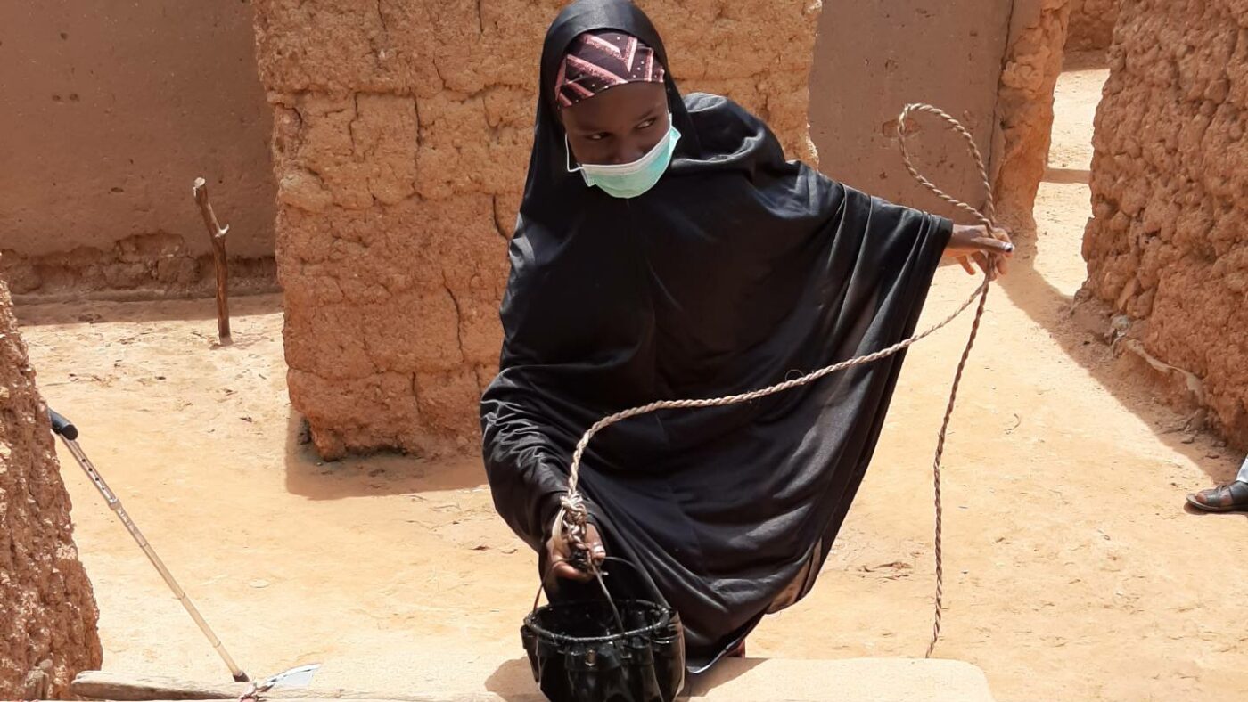 A young woman uses a rope to pull a small vessel of water up from a well.