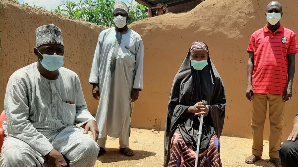 A young woman with a stick sits in the centre of the photo with two men standing and two men seated either side of her, all socially distanced and wearing masks for COVID-19