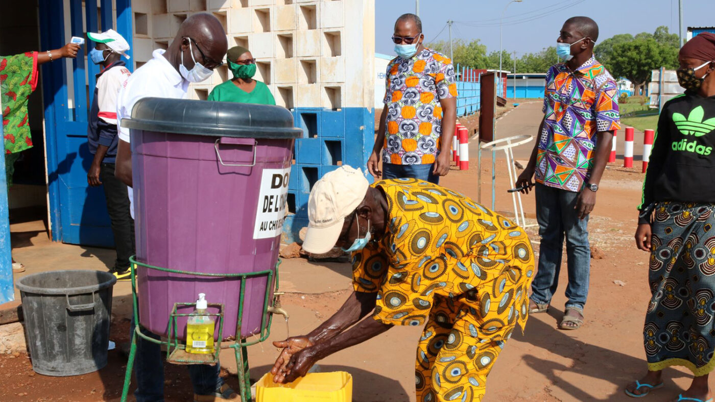 A man washes his hands while wearing a mask, while in the background people wear masks and one has his temperature taken.