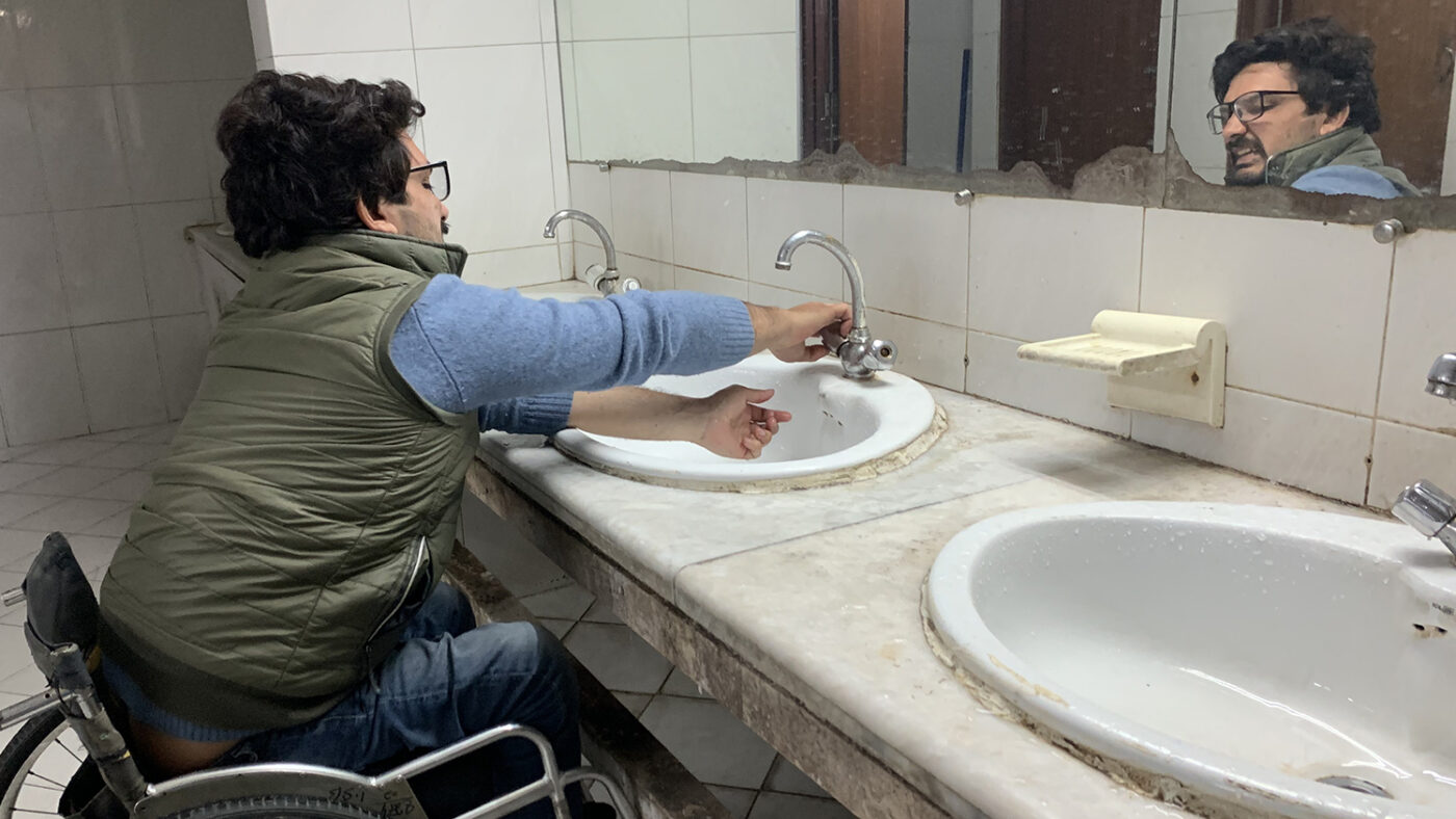A man in a wheelchair reaches up to a sink to turn on the tap.
