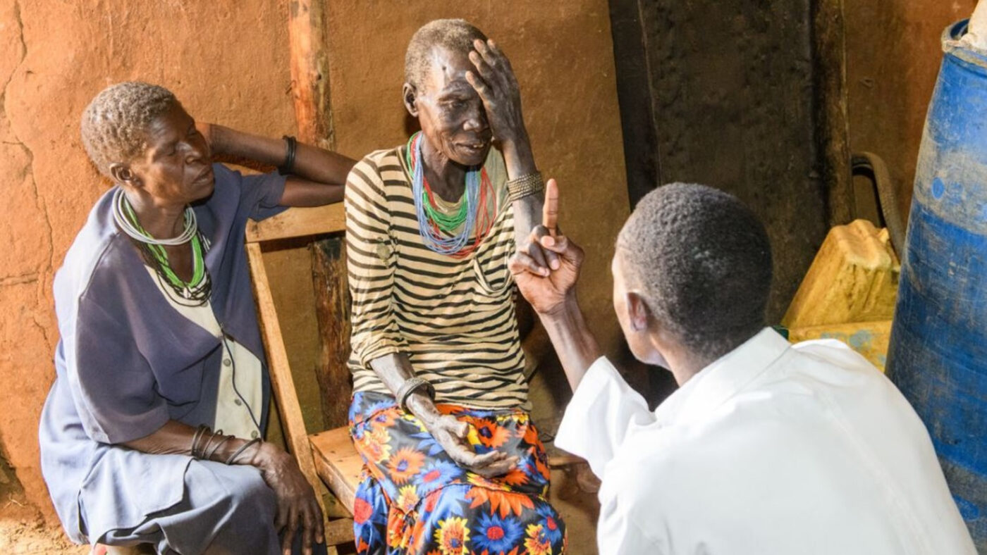 An elderly woman sits in a chair, and receives an eye examination from a man wearing a white shirt. Another woman wearing a blue nurse's uniform sits next to her.