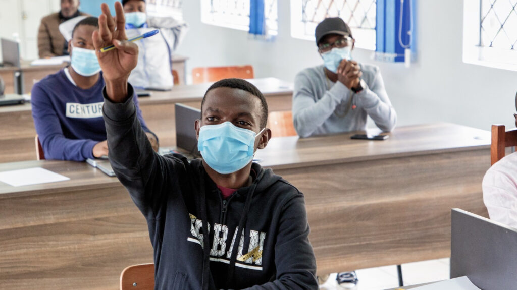 A man at a desk in a classroom raising his hand.