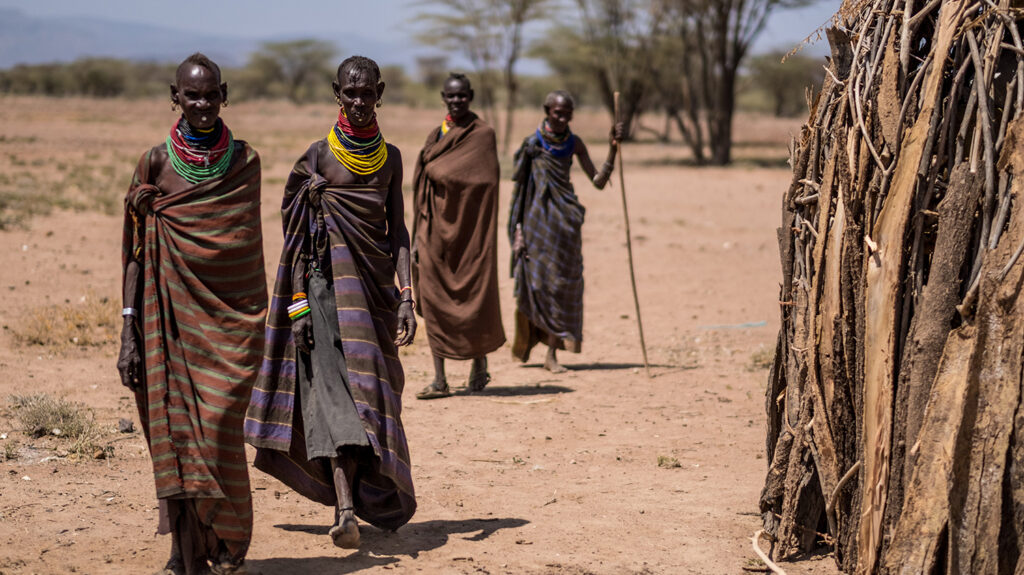 Four adults wearing brightly coloured beaded necklaces are walking outside in pairs. One person is using a stick to assist them.