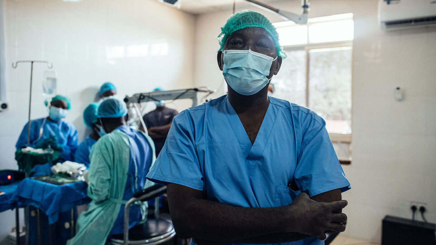 A surgeon in full scrubs stands in the foreground while his team operate behind him.