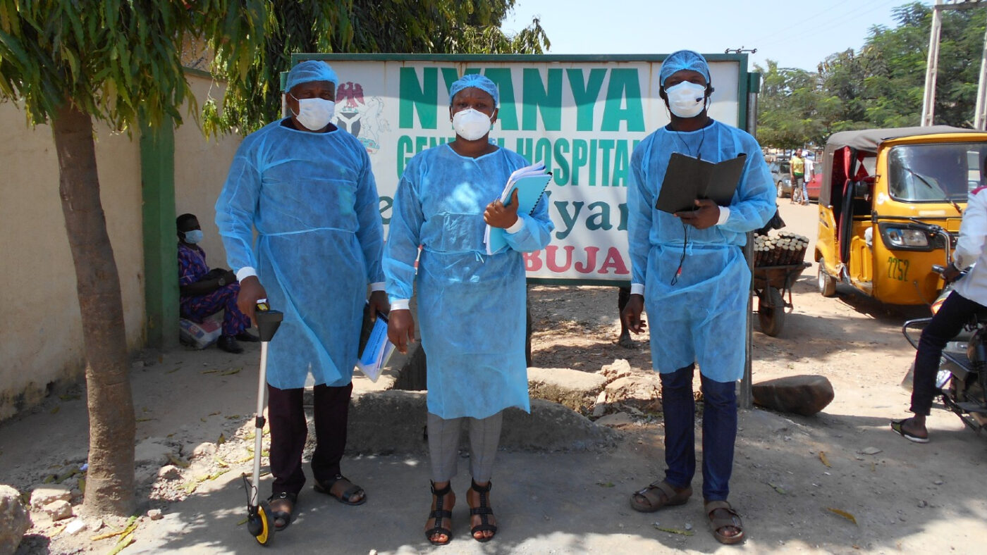 Three people in protective clothing and masks stand outside conducting Covid research.