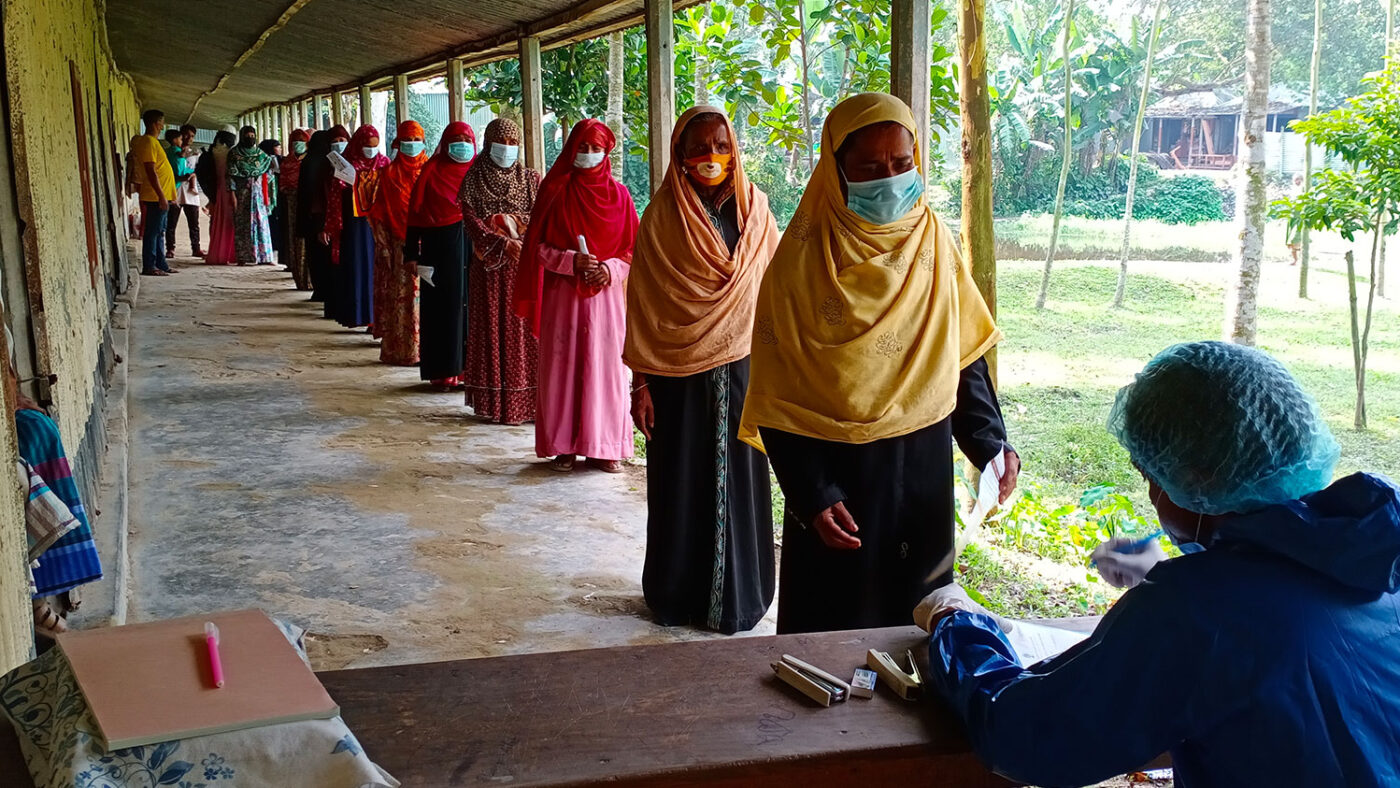 Patients wearing face masks form a social distanced queue in front of a desk.