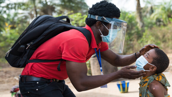 An eye health worker wearing a mask and face shield checks a child's eyes for signs of eye disease.