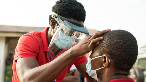 An eye health worker wearing a mask and visor checks a man's eyes for signs of trachoma.