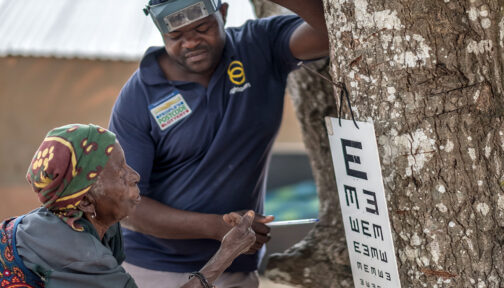 An eye health worker wearing a navy t-shirt points to an eye test chart hanging on a tree, while an older woman looks at the chart.