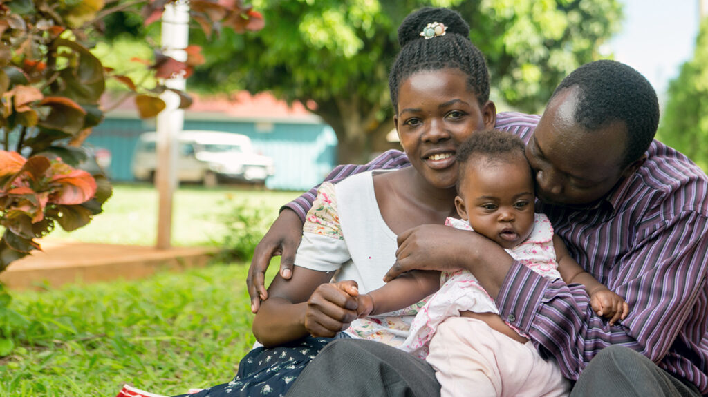 A husband, wife and child sit on the grass embracing amid lush green trees.