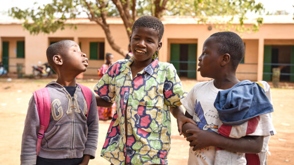 Three boys, one with visual impairments, smile and link arms in their school playground in Mali.
