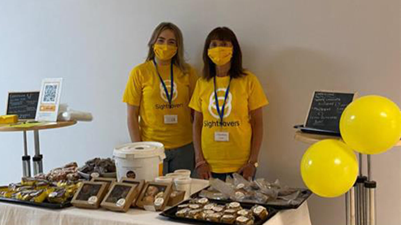 Two people wearing masks and yellow outfits standing in front of a table of cakes.