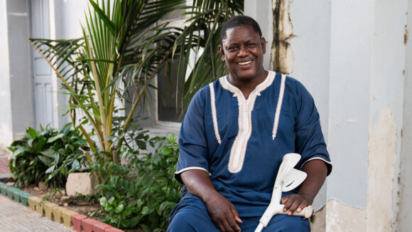 A man smiles at the camera. He's sitting in a chair and holding a white crutch.