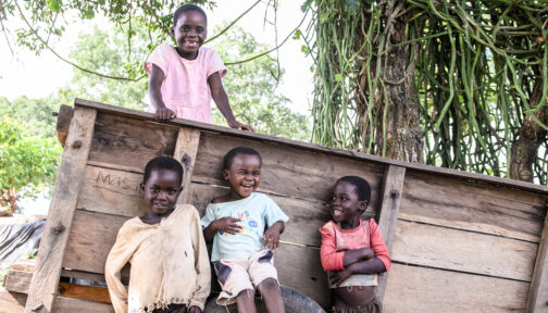 Four children smile and laugh as they sit on a wooden cart amid lush green trees.