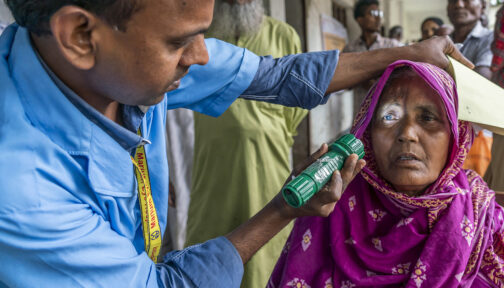 An eye health worker shines a torch into a woman's eyes to check for eye issues. She's wearing a colourful pink headscarf.