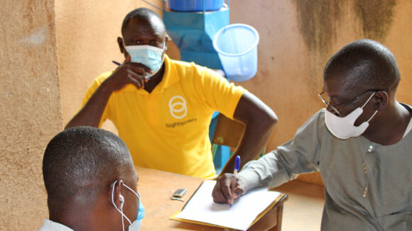 Three people, one in a yellow Sightsavers t-shirt, sit round a table filling in paperwork.