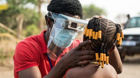 An eye health worker wearing a mask and visor checks a child's eyes for signs of trachoma.