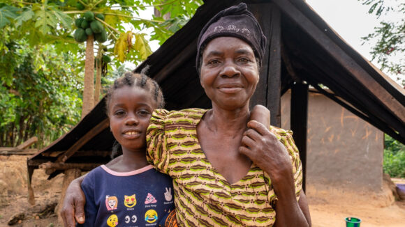 Angelyn and her grandmother, link arms and smile, with dusty ground and trees in the background.
