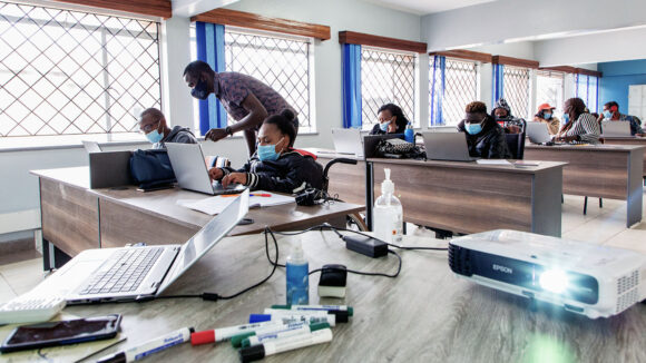 Students sit in front of laptops at desks in a classroom. In the foreground, a teacher stands next to a student, giving instruction.